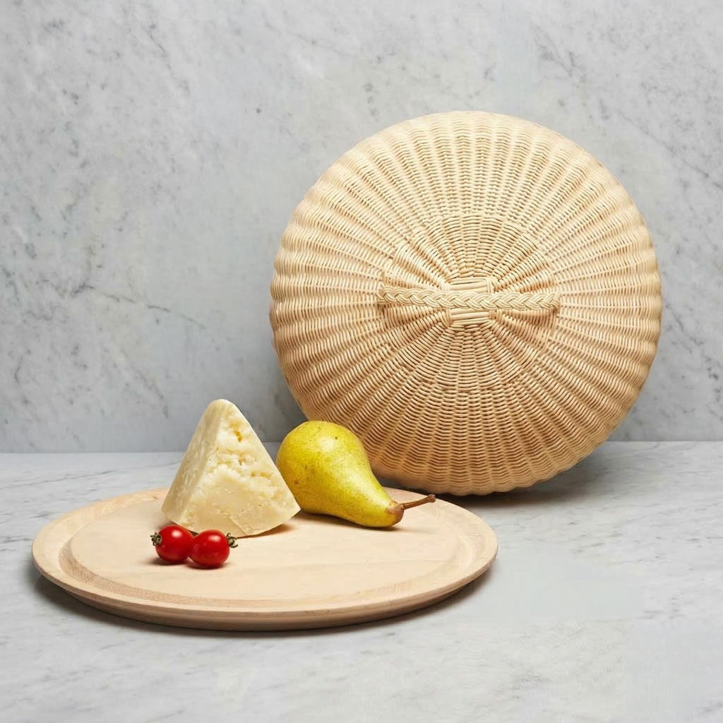 Round light wood serving plate displaying a wedge of hard cheese, a yellow pear, and two cherry tomatoes with a large woven wicker dome cover with handle.
